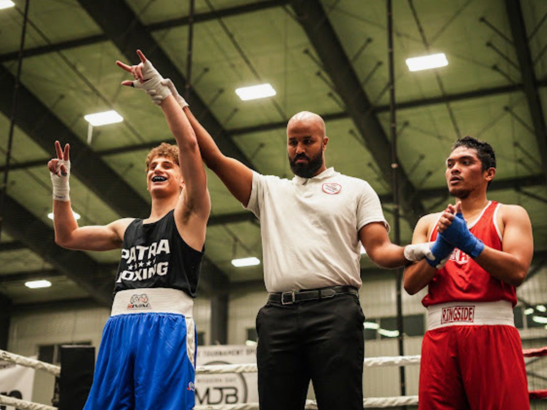 A Patra Boxing athlete in blue has his hand raised by the referee after a bout.